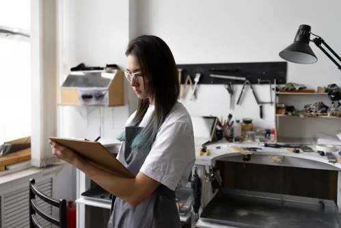 Young woman Jeweler in goggles marks in her clipboard completed orders for manufacture of products. taller